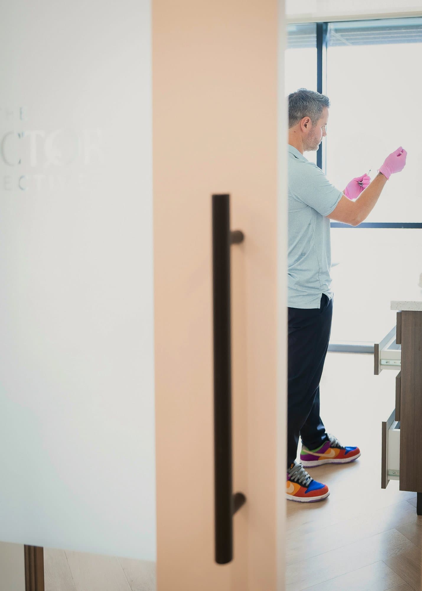 Ben Tracy, MD, seen through a frosted clinic door, prepping a syringe in very colourful sneakers.