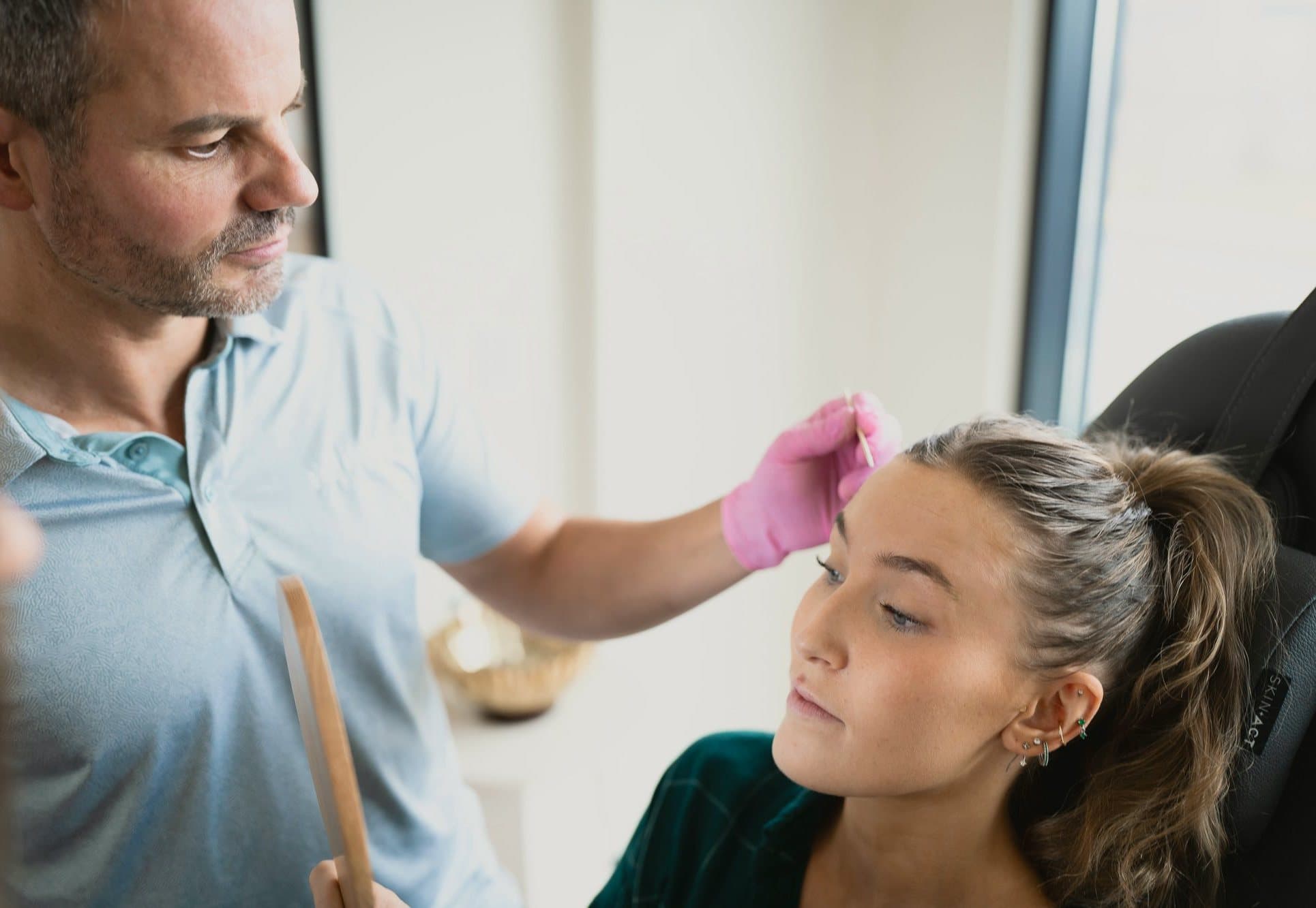 Ben Tracy, MD, in a pale blue polo and pink gloves, mapping a patient's brow in a bright clinic room.